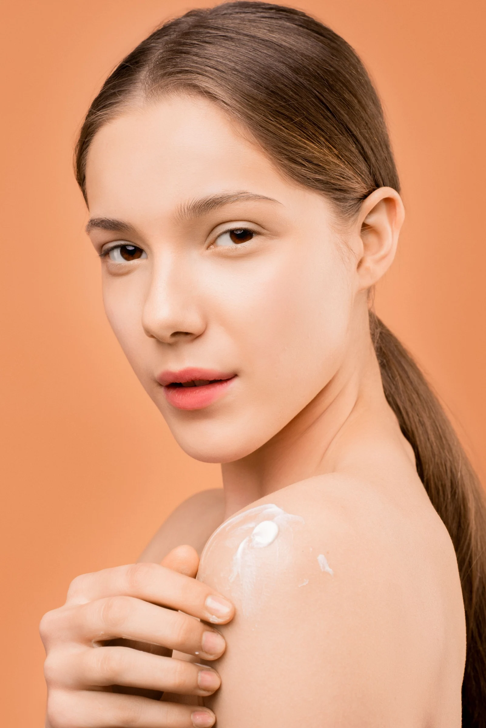 A woman gently applying moisturiser to her shoulder against a warm peach background